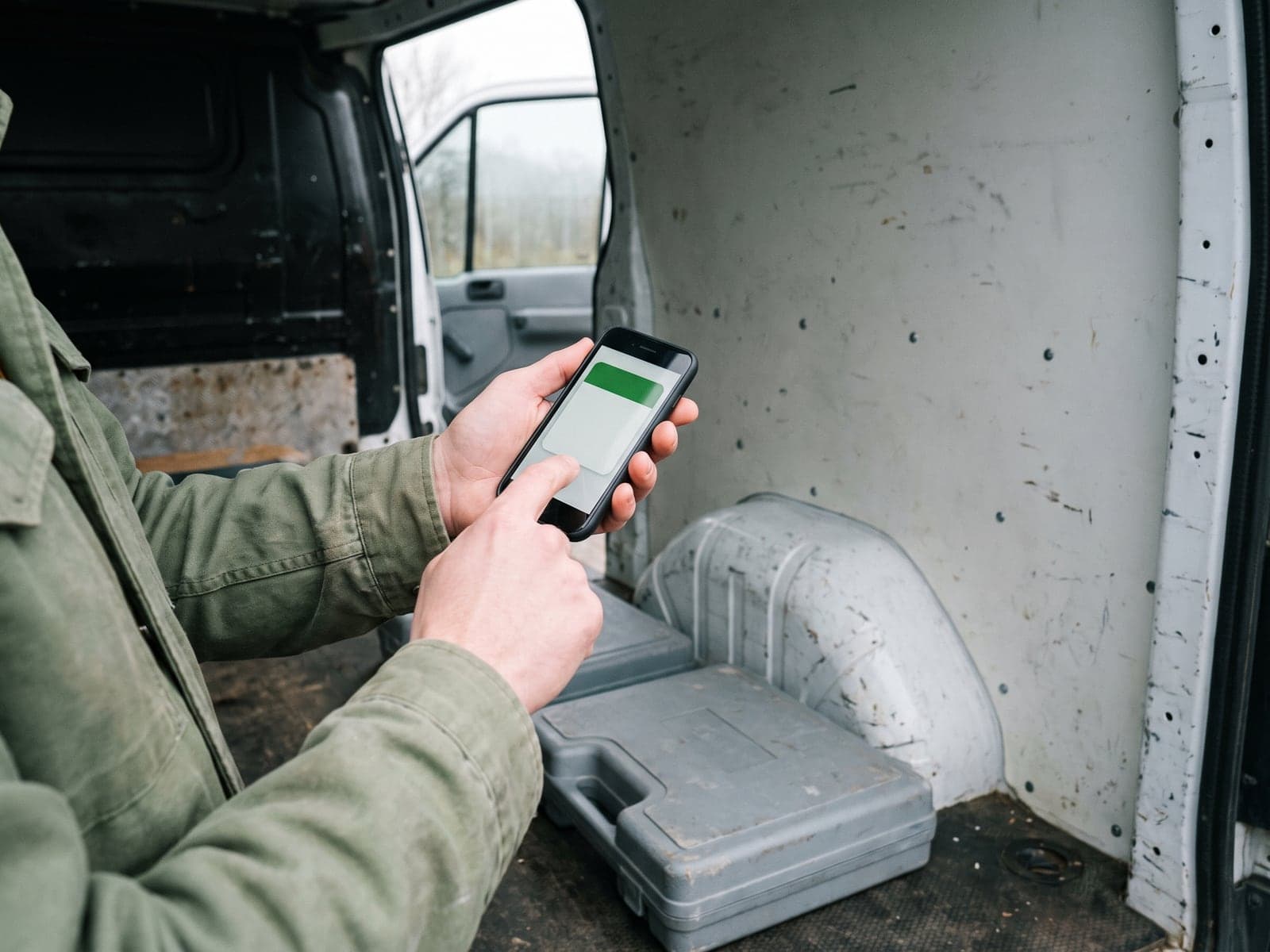 A tradesperson in an olive work jacket taps a job card on a phone inside the cargo area of a van.