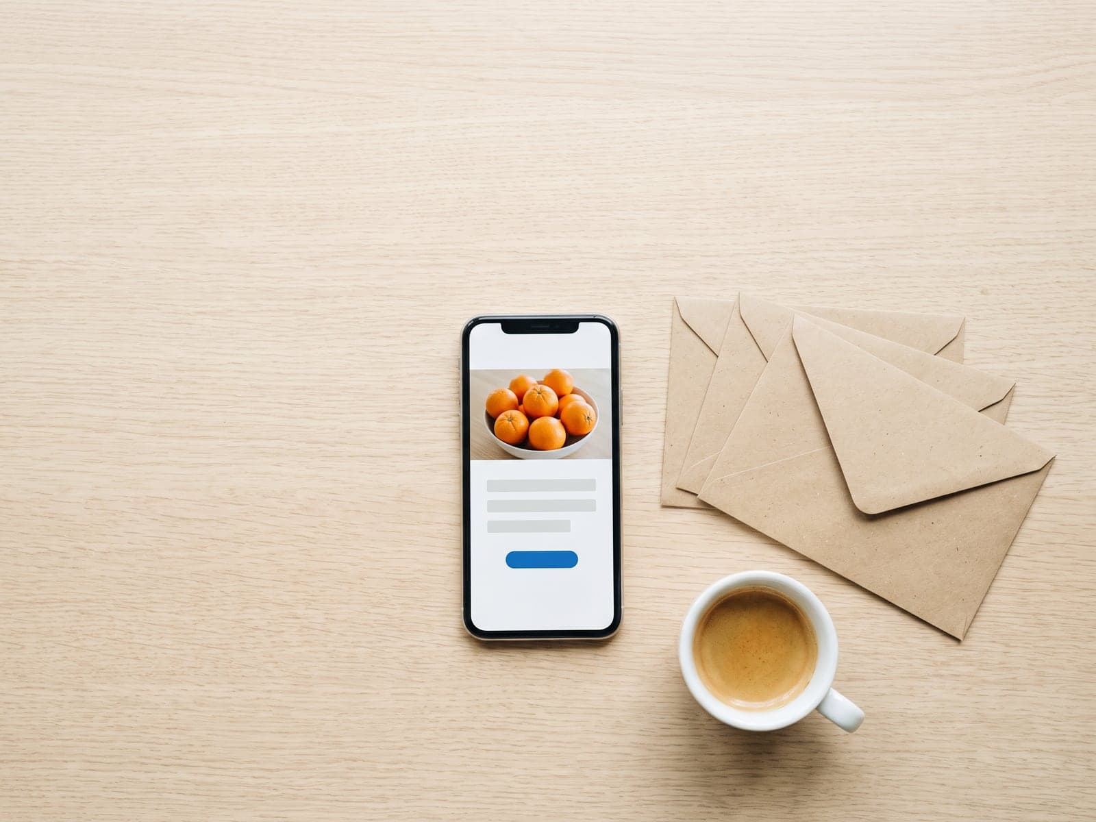 A phone showing an email with a photo of oranges and a blue button lies on a desk beside kraft envelopes and an espresso.