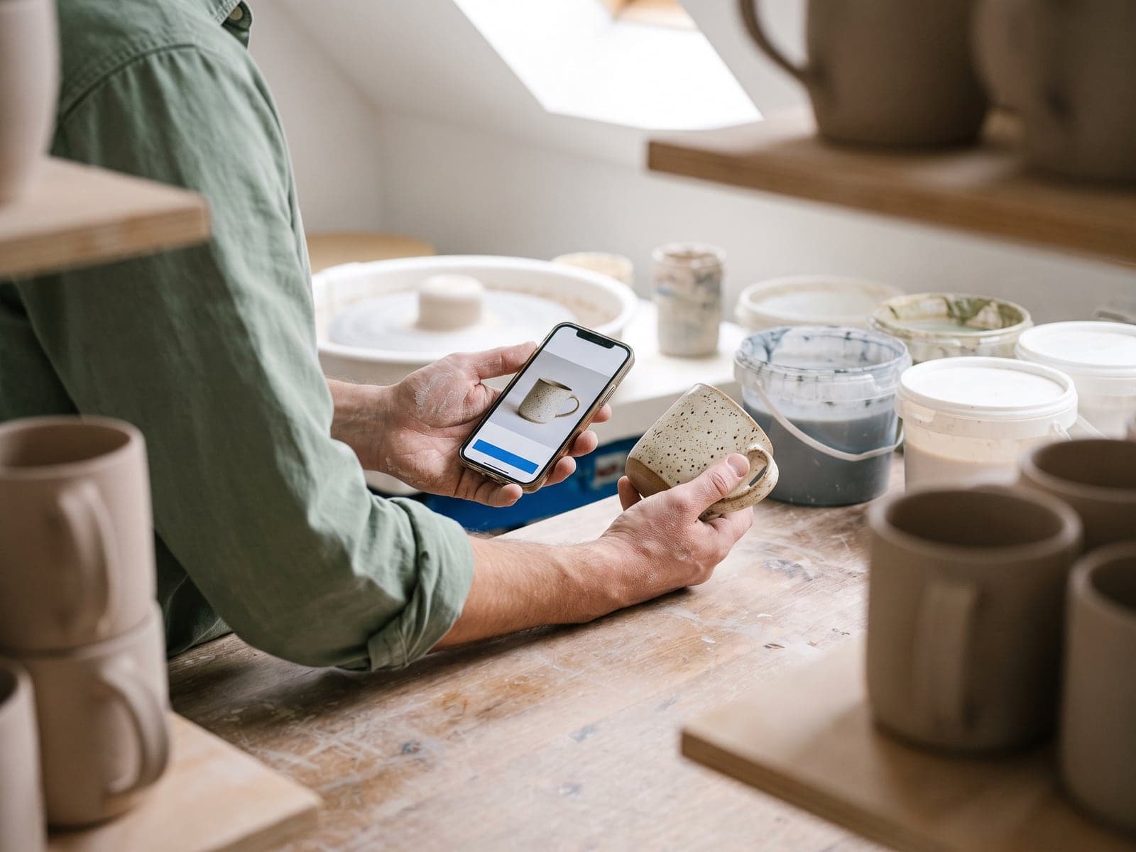 A potter's clay-dusted hands hold a phone showing an ad for the speckled mug held in the other hand.