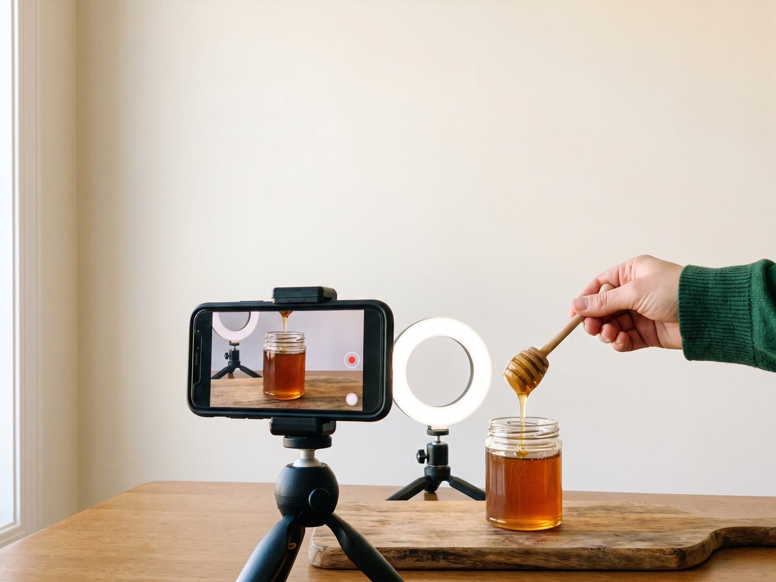 A phone on a mini tripod films honey dripping from a wooden dipper into a jar, lit by a small ring light.