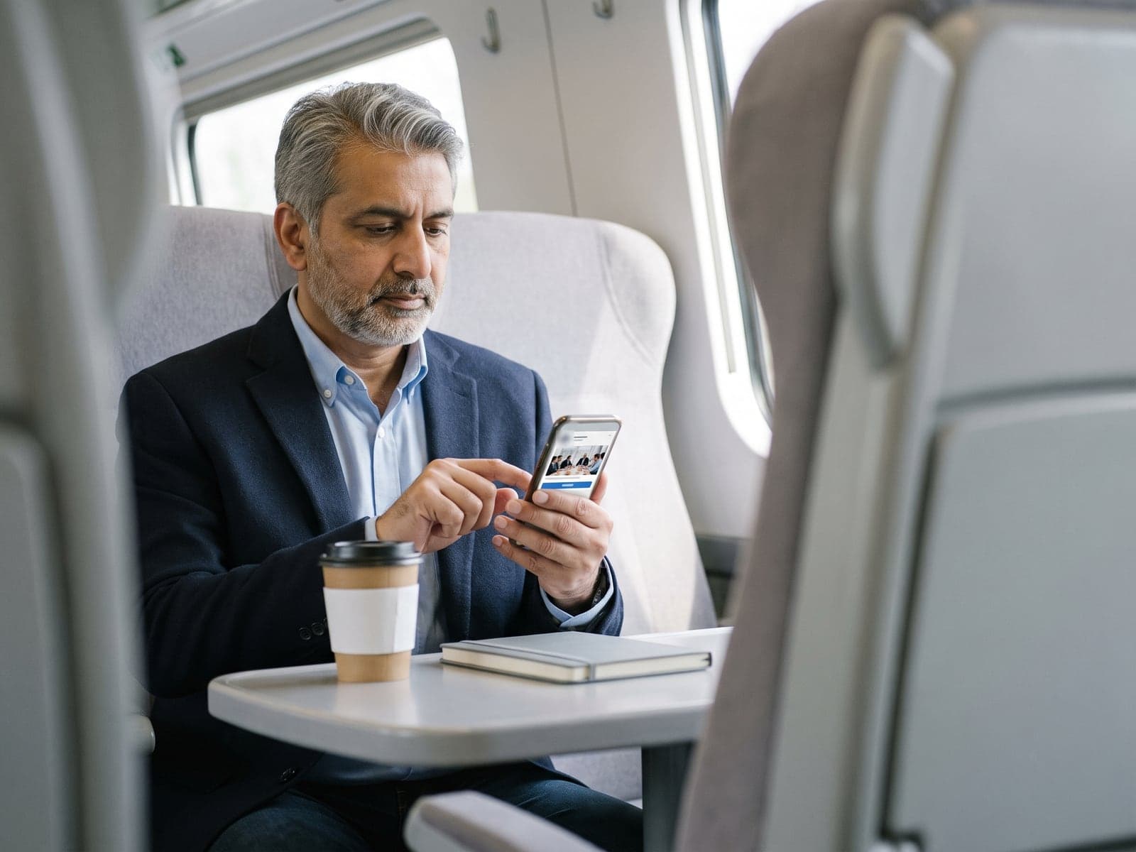 A businessman in a navy blazer scrolls a professional feed on his phone at a table in a bright train carriage.