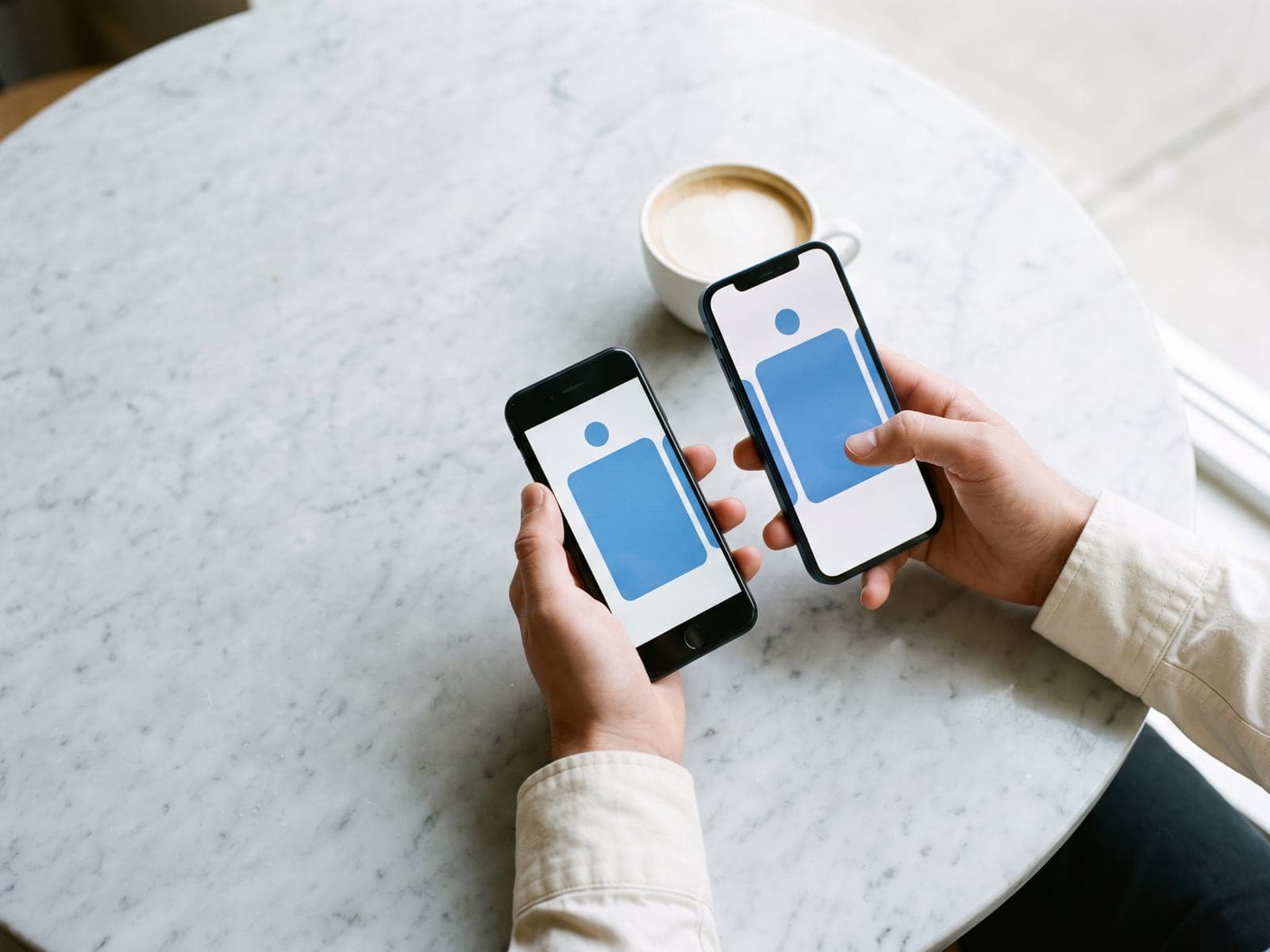 Two hands hold two different phones over a round marble cafe table, both showing the same blue app card.