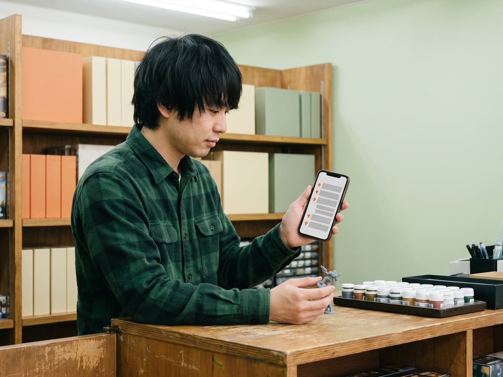 A hobby shop owner holds a miniature figure while his phone shows a discussion thread with orange upvote arrows.
