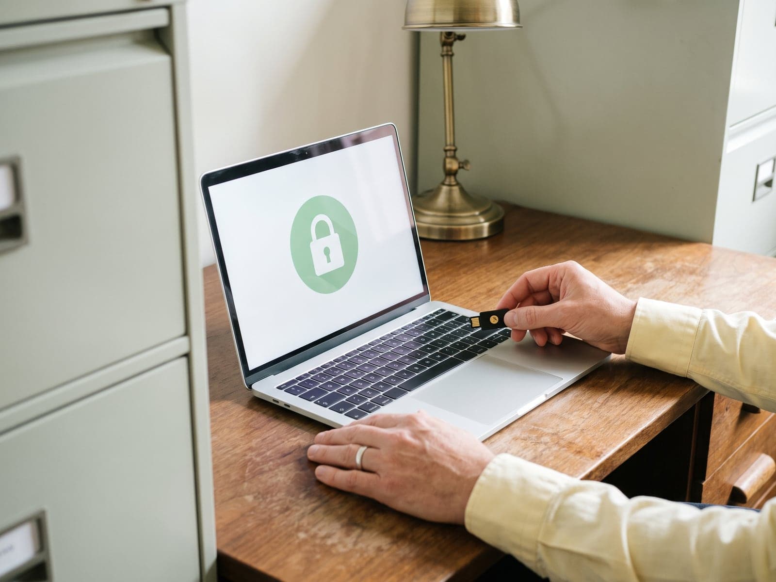 A hand holds a hardware security key over a laptop showing a green padlock in a small office with sage filing cabinets.