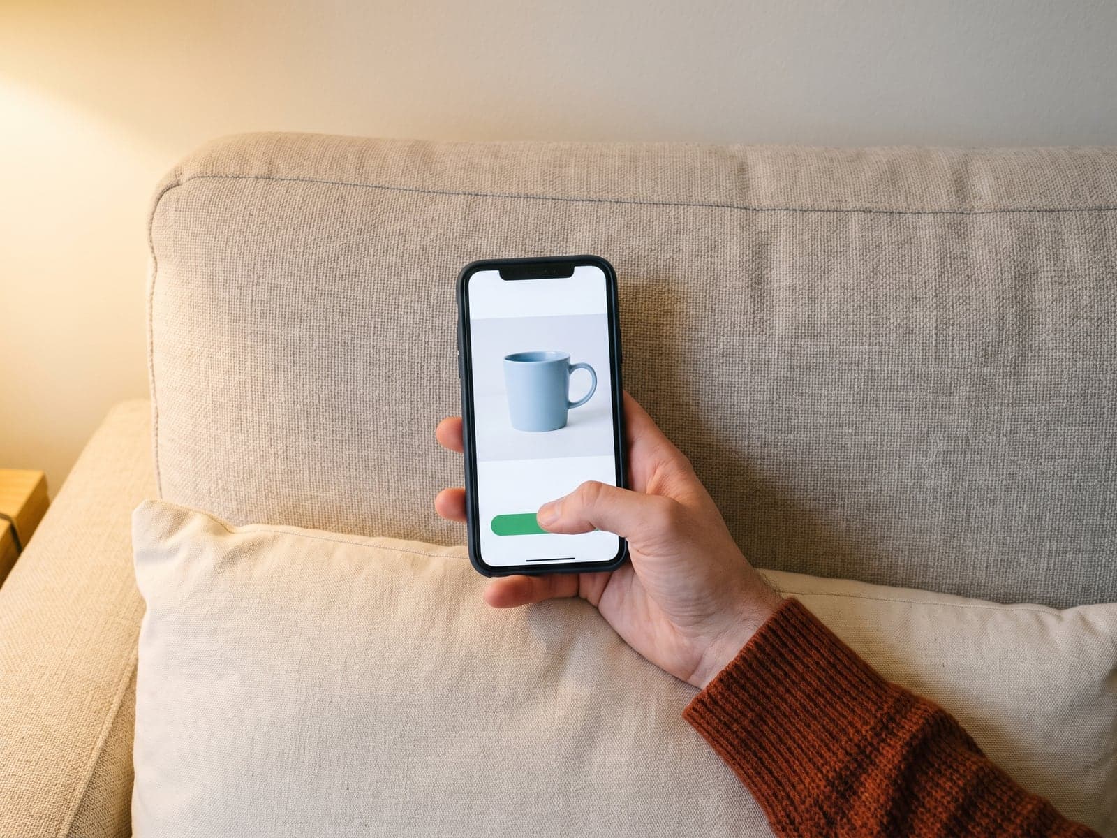 A hand holds a phone over a sofa cushion, thumb hovering over a green checkout button beneath a photo of a blue mug.