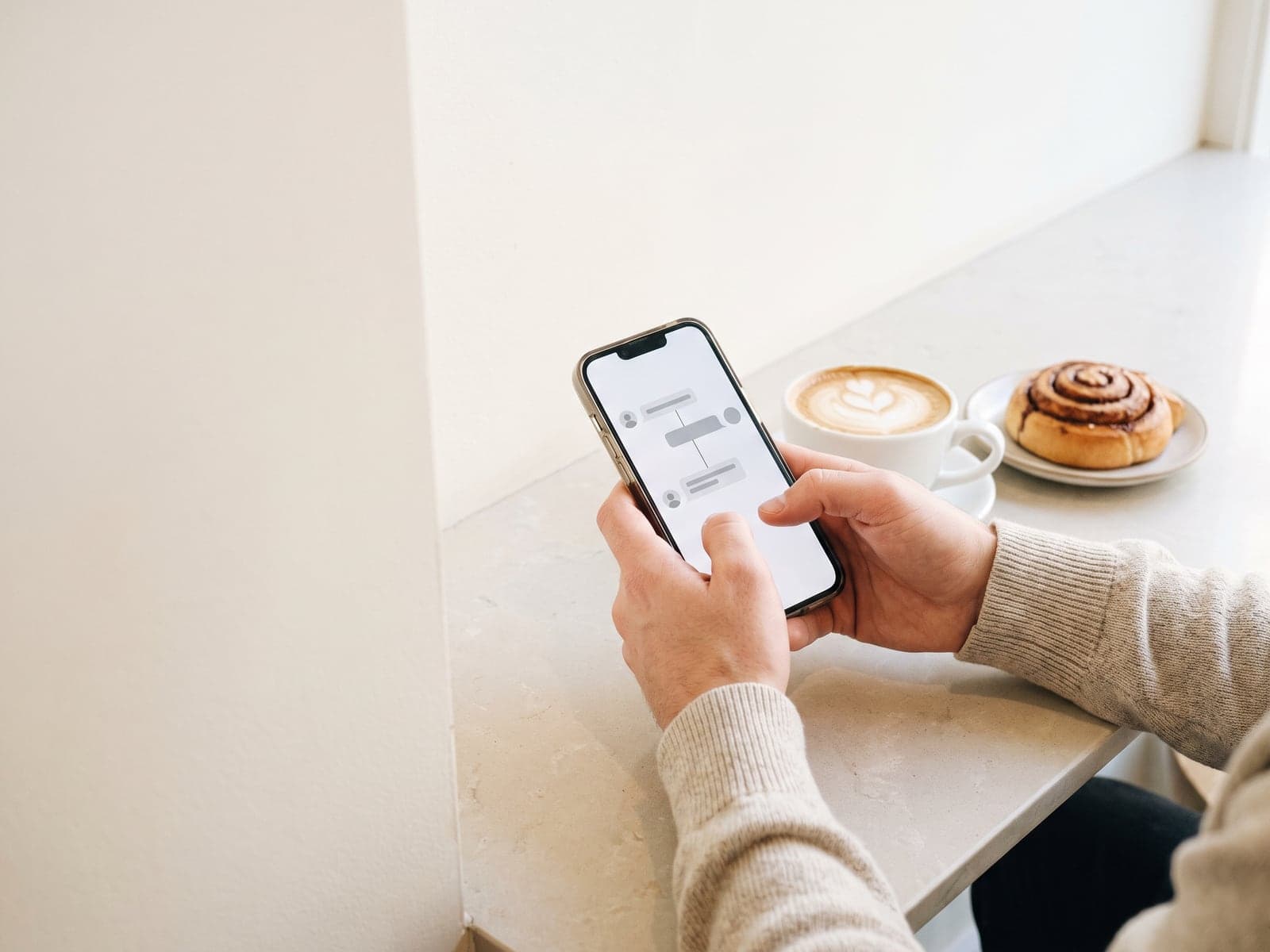 Two hands type a short thread of connected posts on a phone at a bakery cafe counter, with a cappuccino and a cinnamon bun.