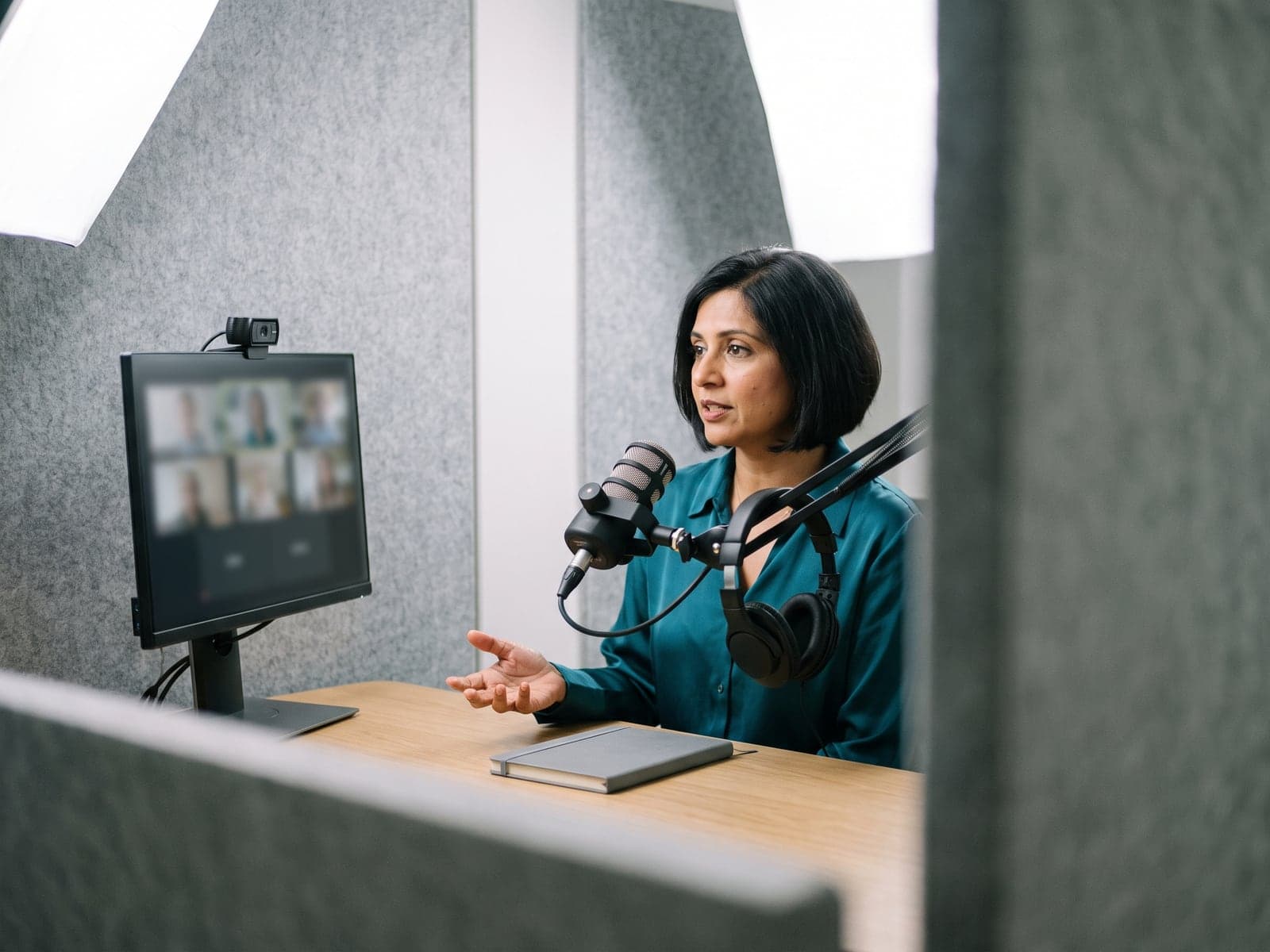 A presenter speaks into a broadcast microphone in a grey felt webinar booth while a monitor shows a grid of attendees.