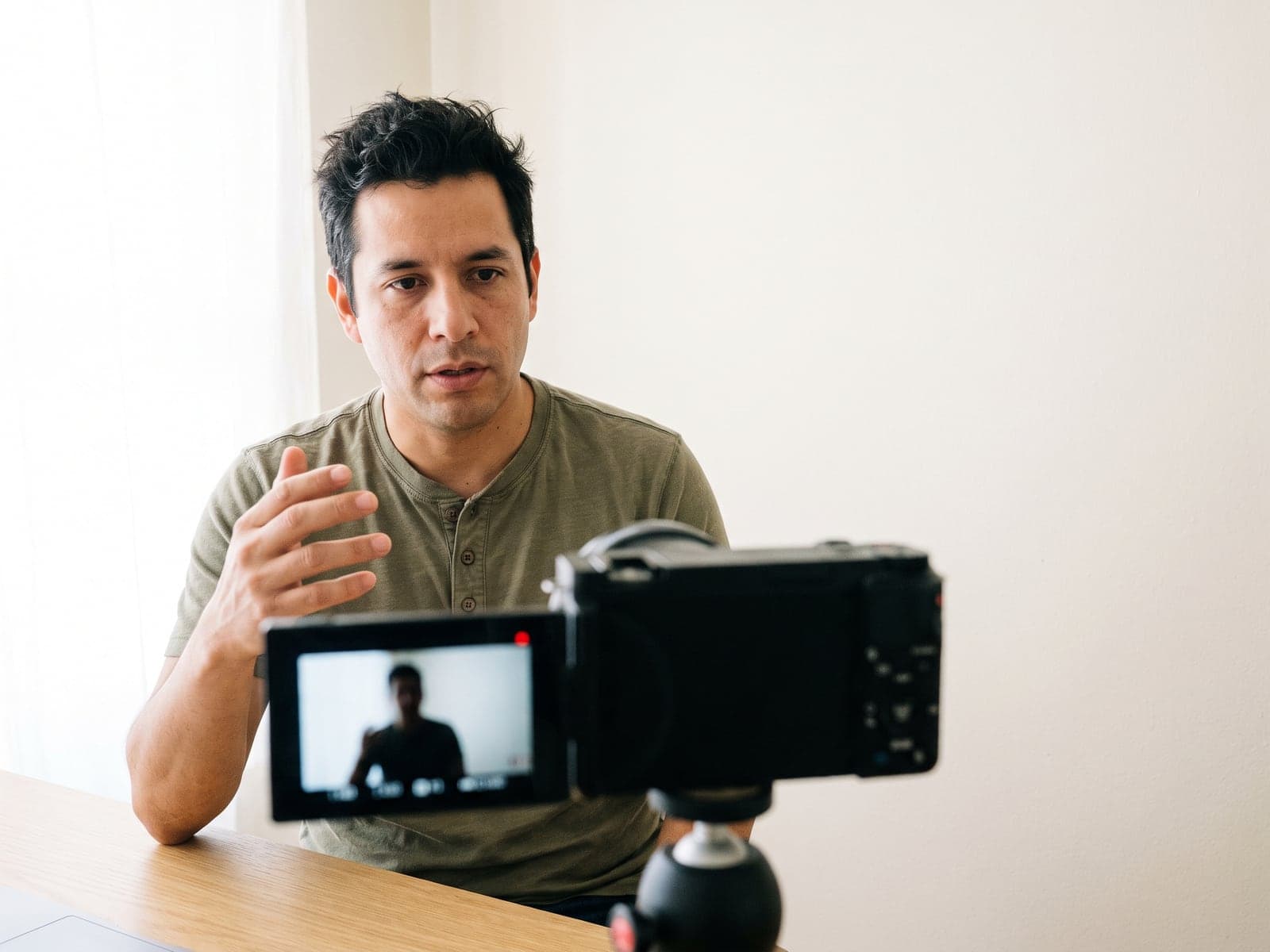 A creator talks to a camera on a tripod while recording a video at his desk in a bright home studio.